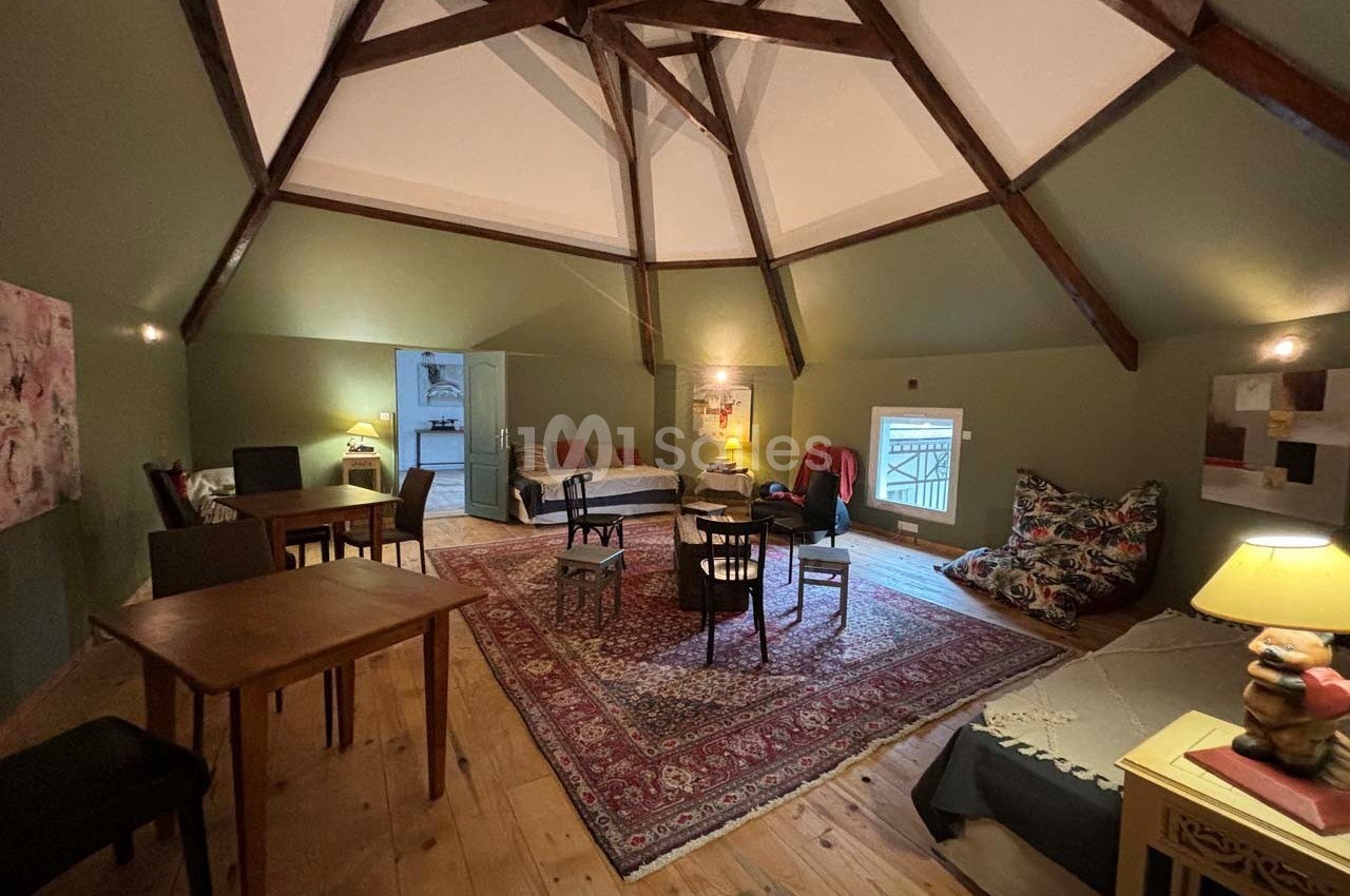 Salle mansardée avec poutres apparentes, tapis au sol, mobilier varié et éclairage chaleureux.