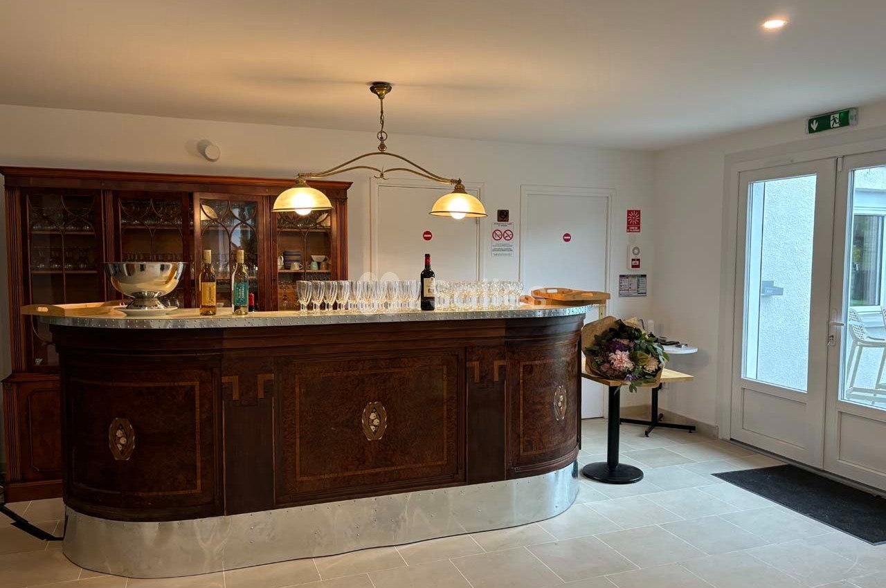 Comptoir en bois ancien avec des verres alignés, une bouteille de vin et une décoration florale à côté.
