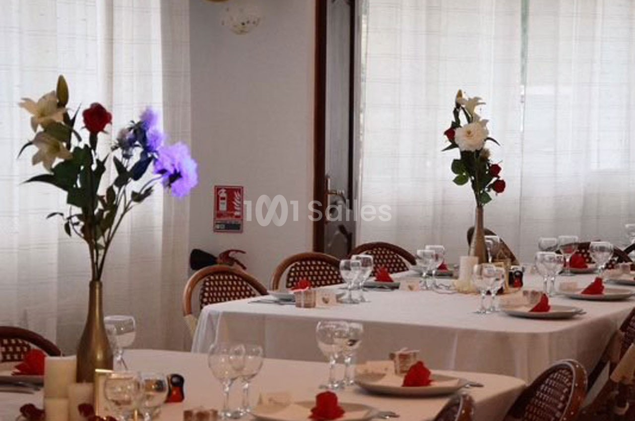 Salle de réception avec tables dressées, nappes blanches, verres, serviettes rouges et vases de fleurs.