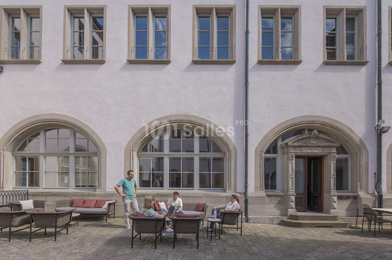Personnes assises et discutant dans une cour pavée devant un bâtiment aux grandes fenêtres et arches.