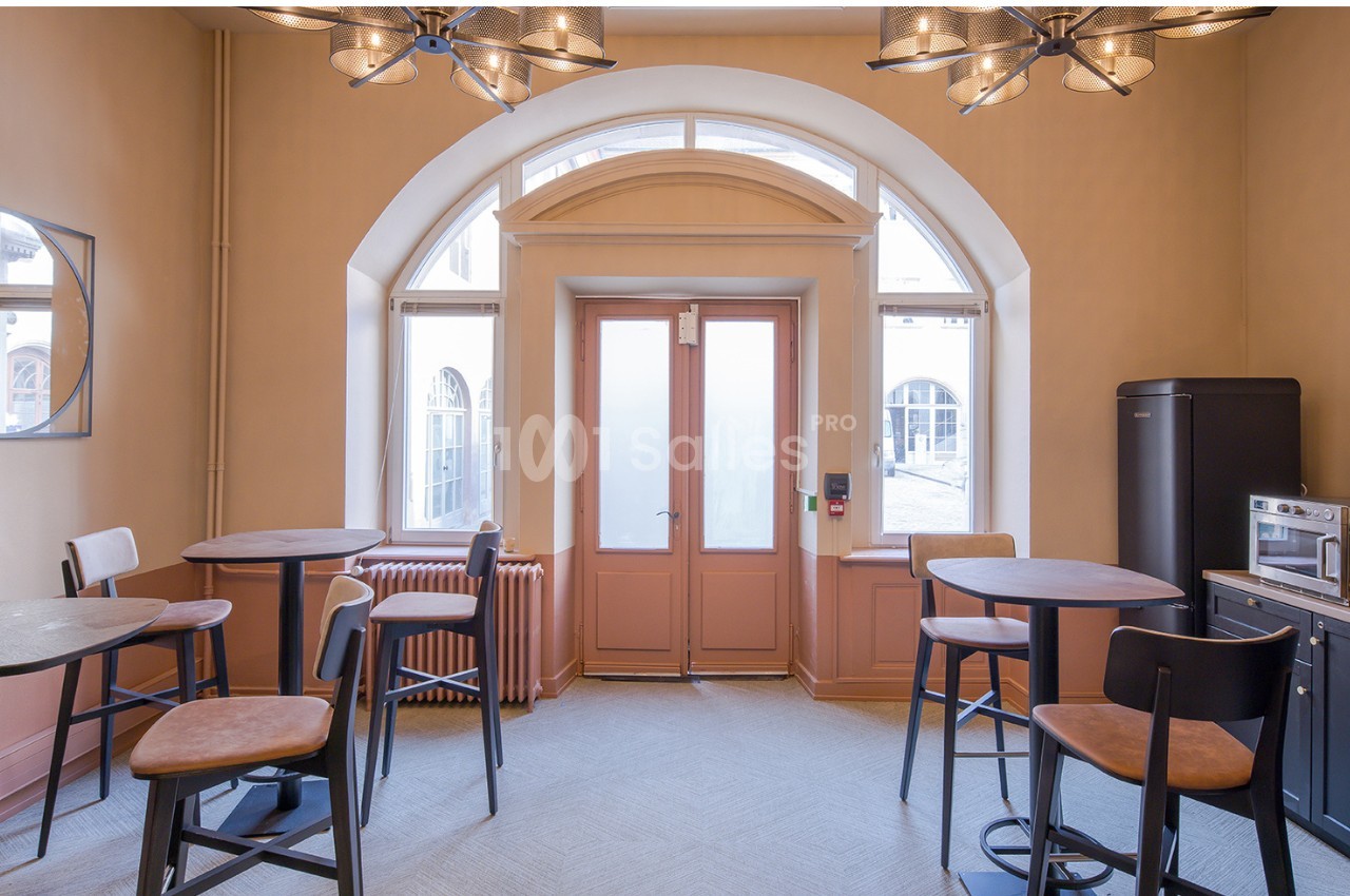 Salle lumineuse avec tables hautes, chaises, grande porte vitrée, fenêtres cintrées et coin kitchenette équipé.