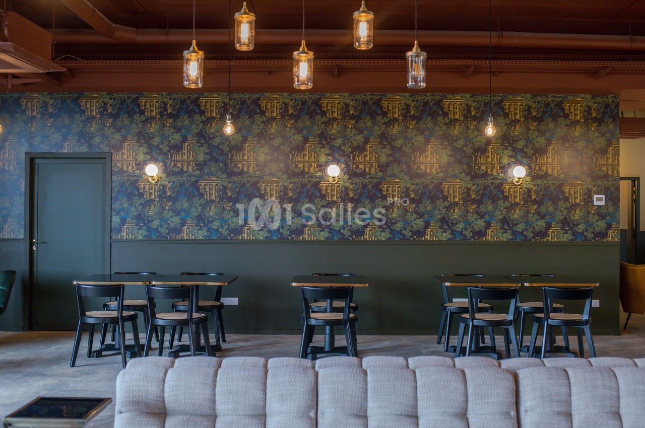 Salle de restaurant avec tables en bois, chaises noires, mur décoré de papier peint floral et luminaires suspendus.