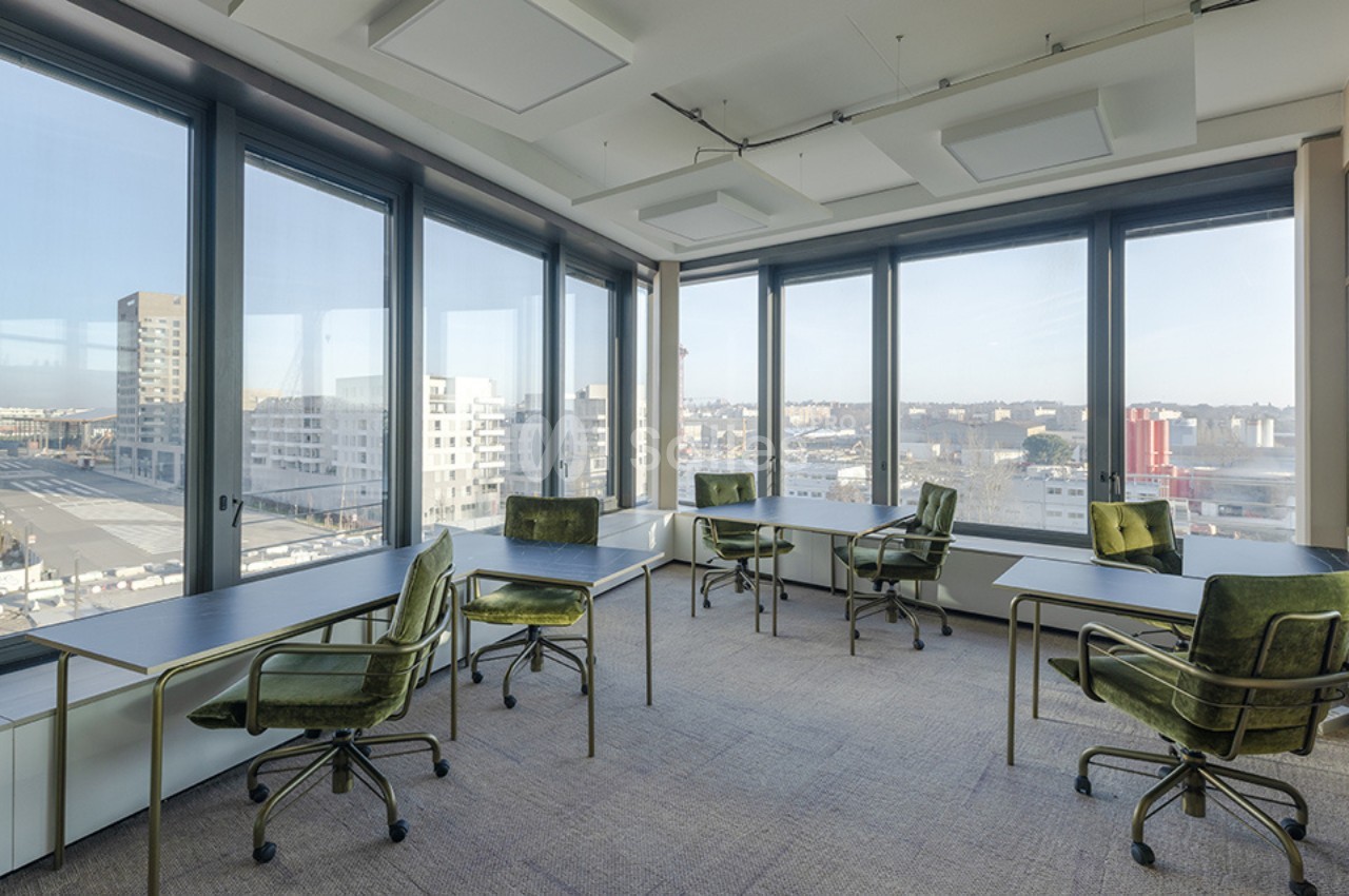 Salle de travail lumineuse avec de grandes baies vitrées, des bureaux espacés et des chaises vertes pivotantes.