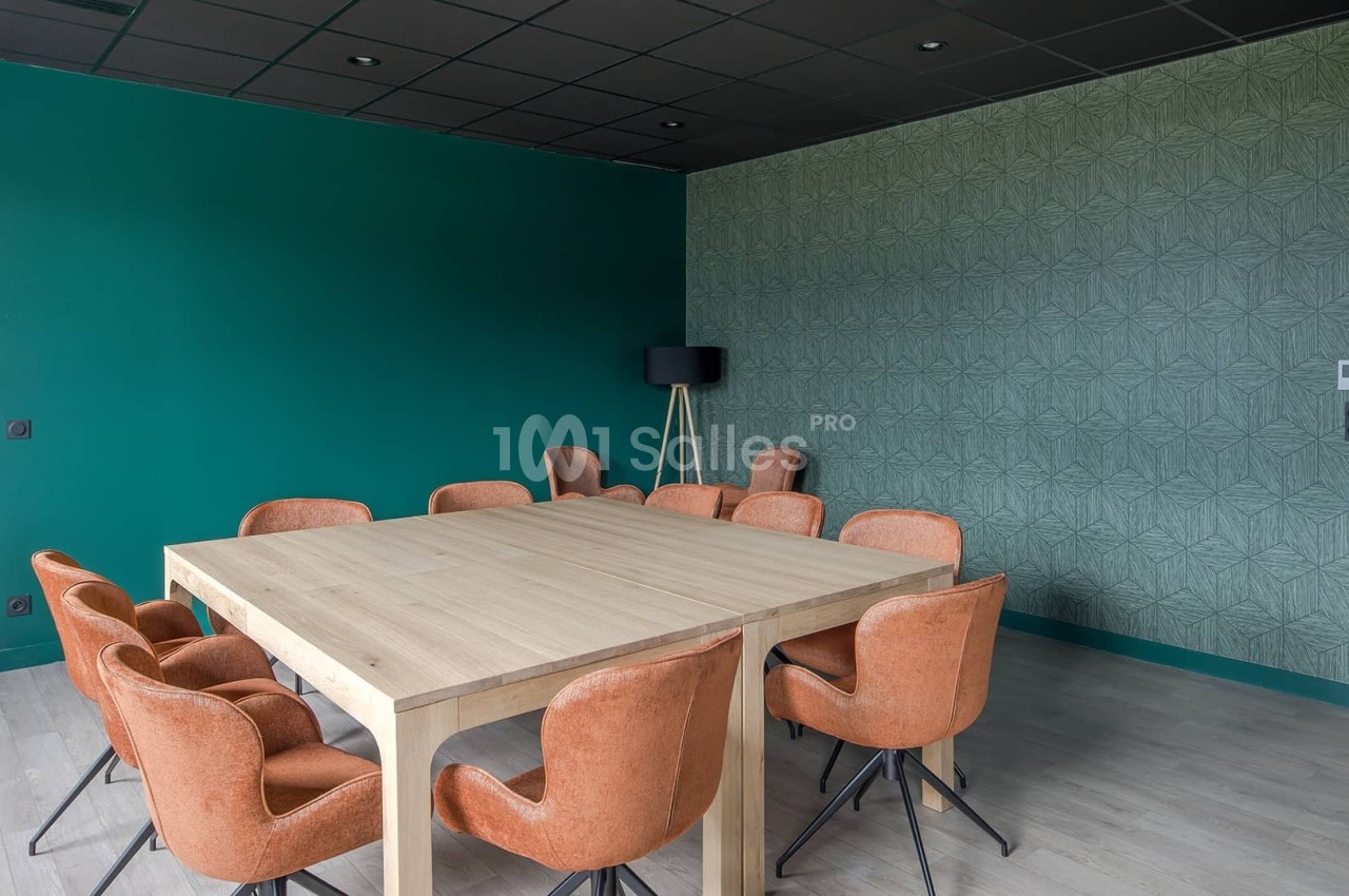 Salle de réunion avec table en bois clair, chaises orange, murs verts et éclairage naturel.