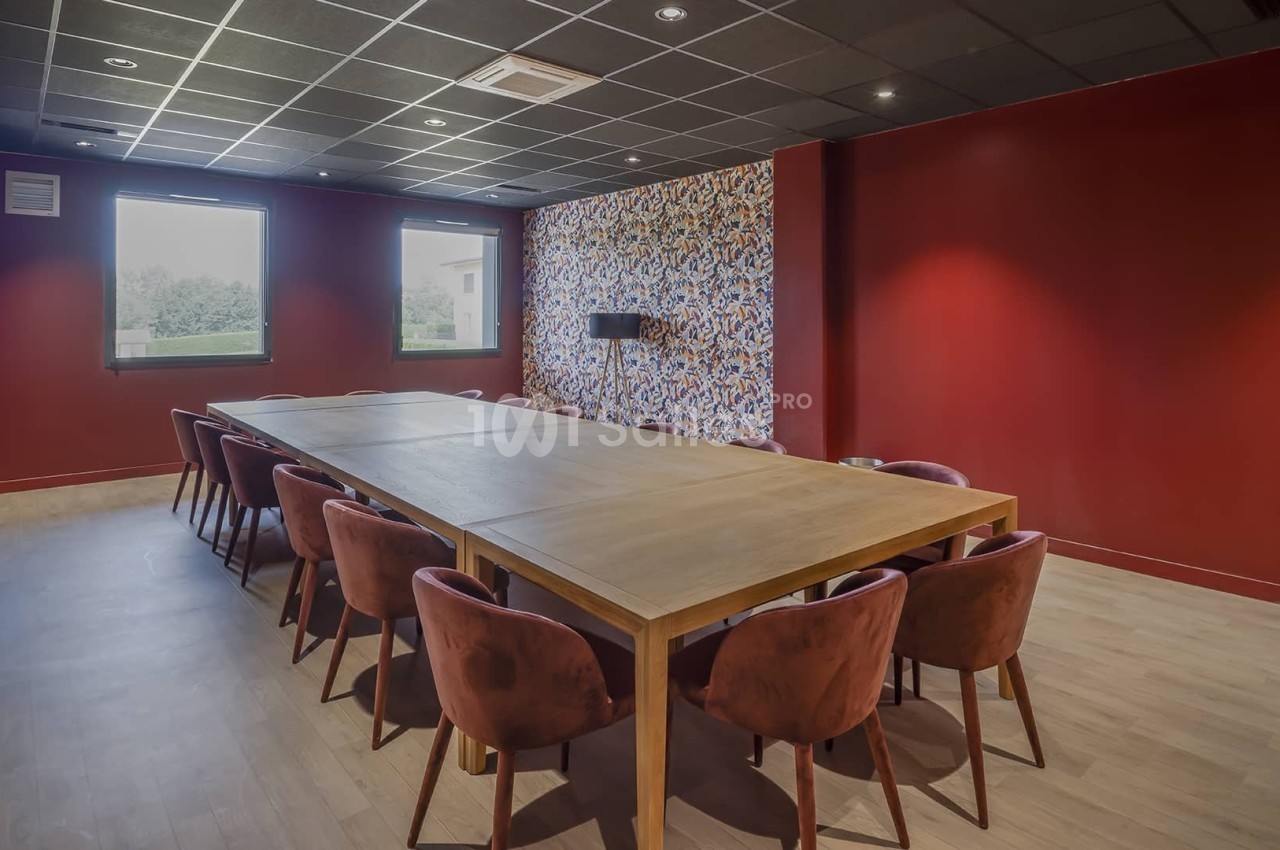 Salle de réunion avec murs rouges, grande table en bois entourée de chaises et lumière naturelle provenant des fenêtres.