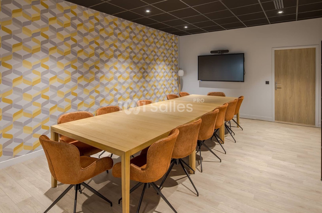 Salle de réunion avec une grande table en bois, des chaises orange, un mur à motifs géométriques et un écran mural.