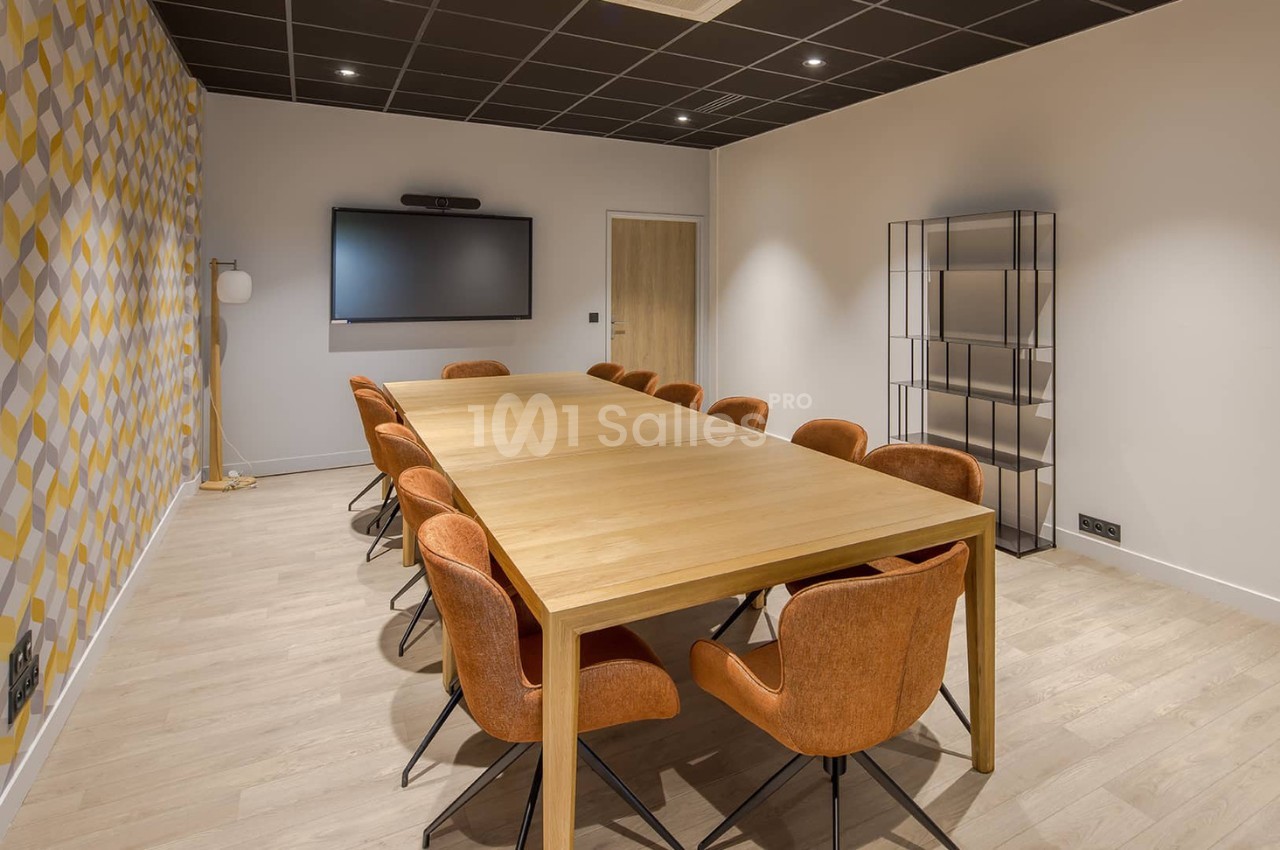 Salle de réunion avec une grande table en bois, chaises orange, écran mural et décoration murale géométrique.