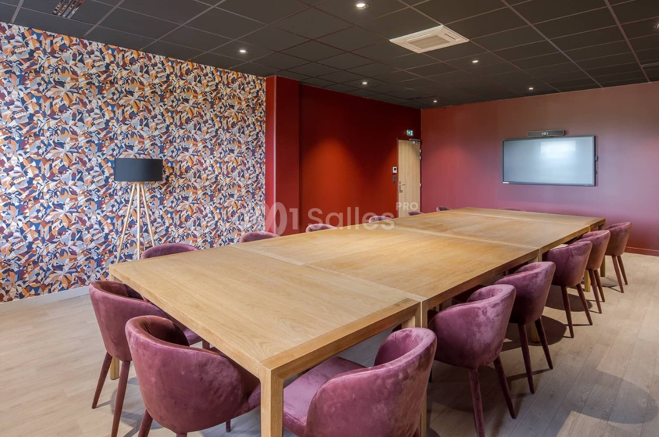 Salle de réunion avec grande table en bois, chaises en velours bordeaux, mur rouge et écran mural.