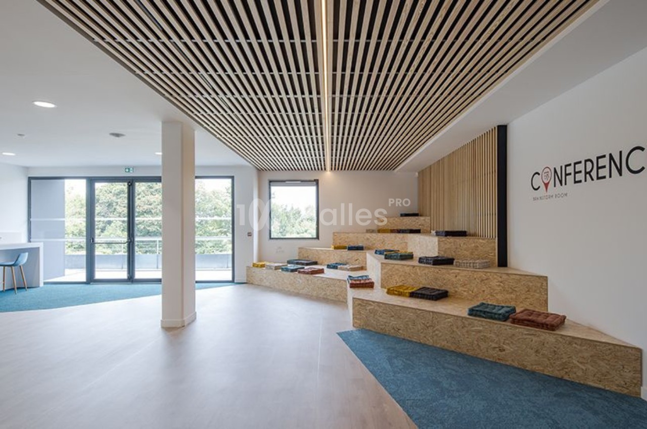 Espace intérieur lumineux avec gradins en bois, coussins colorés, plafond à lattes et grandes fenêtres donnant sur l…