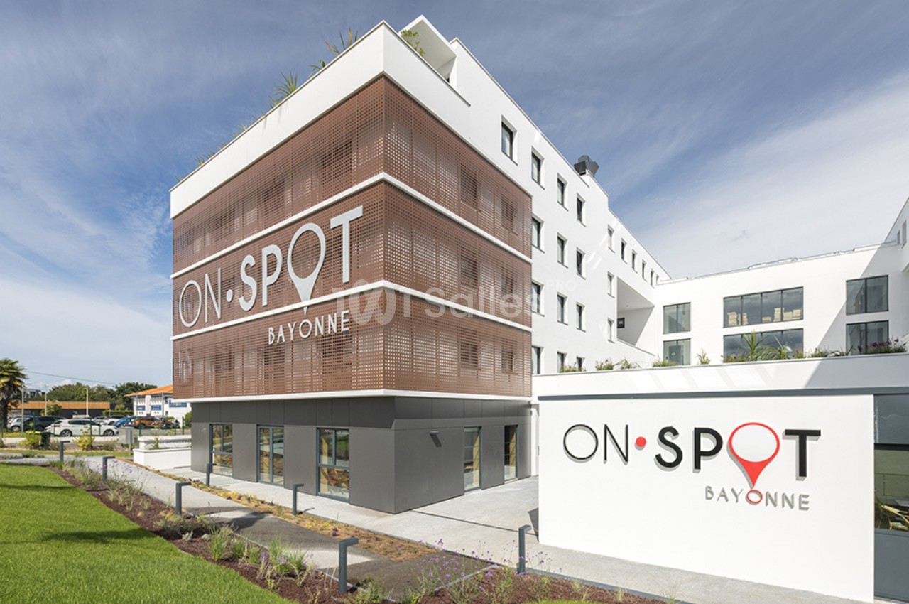 Façade moderne d'un bâtiment blanc avec des panneaux en bois et le logo ’On Spot Bayonne’ visible.