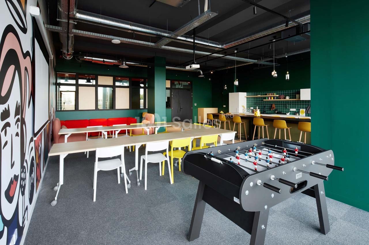 Salle de détente moderne avec baby-foot, tables, chaises colorées et cuisine ouverte en arrière-plan.