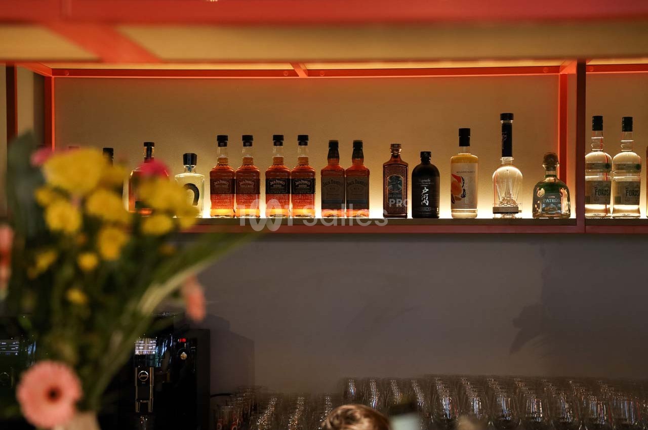 Bouteilles d'alcool alignées sur une étagère rétroéclairée, avec un bouquet de fleurs flou au premier plan.