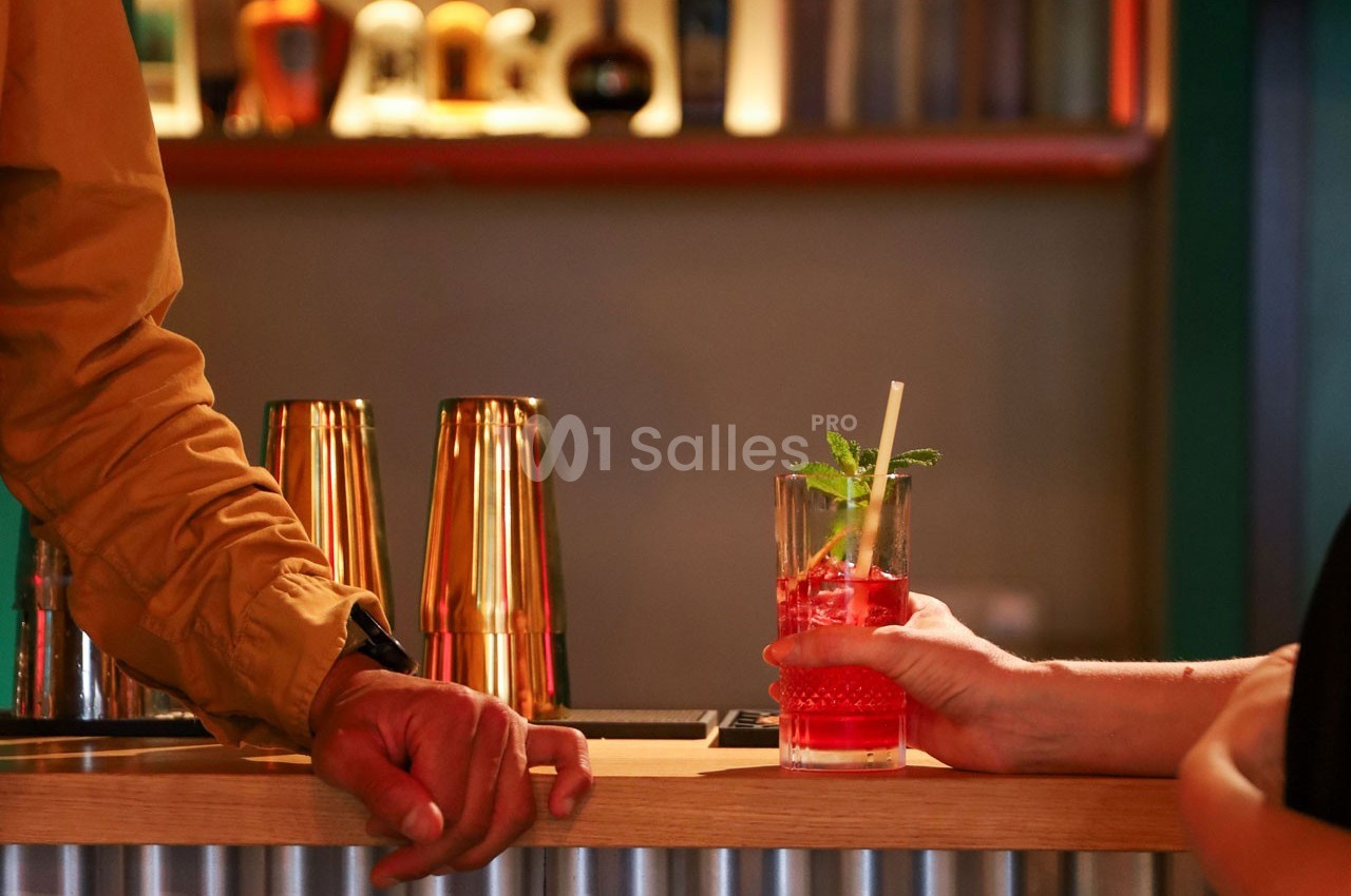Un cocktail rouge garni de menthe est tenu par une main sur un comptoir de bar en bois, avec des shakers en arrière-plan.