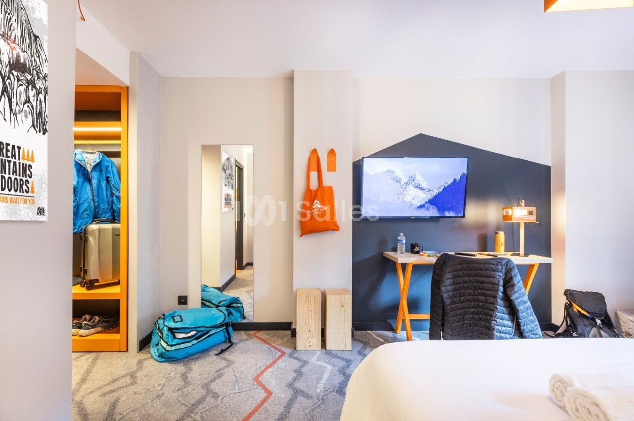 Chambre d'hôtel moderne avec lit, bureau, écran mural, valises et vêtements rangés dans un coin ouvert.