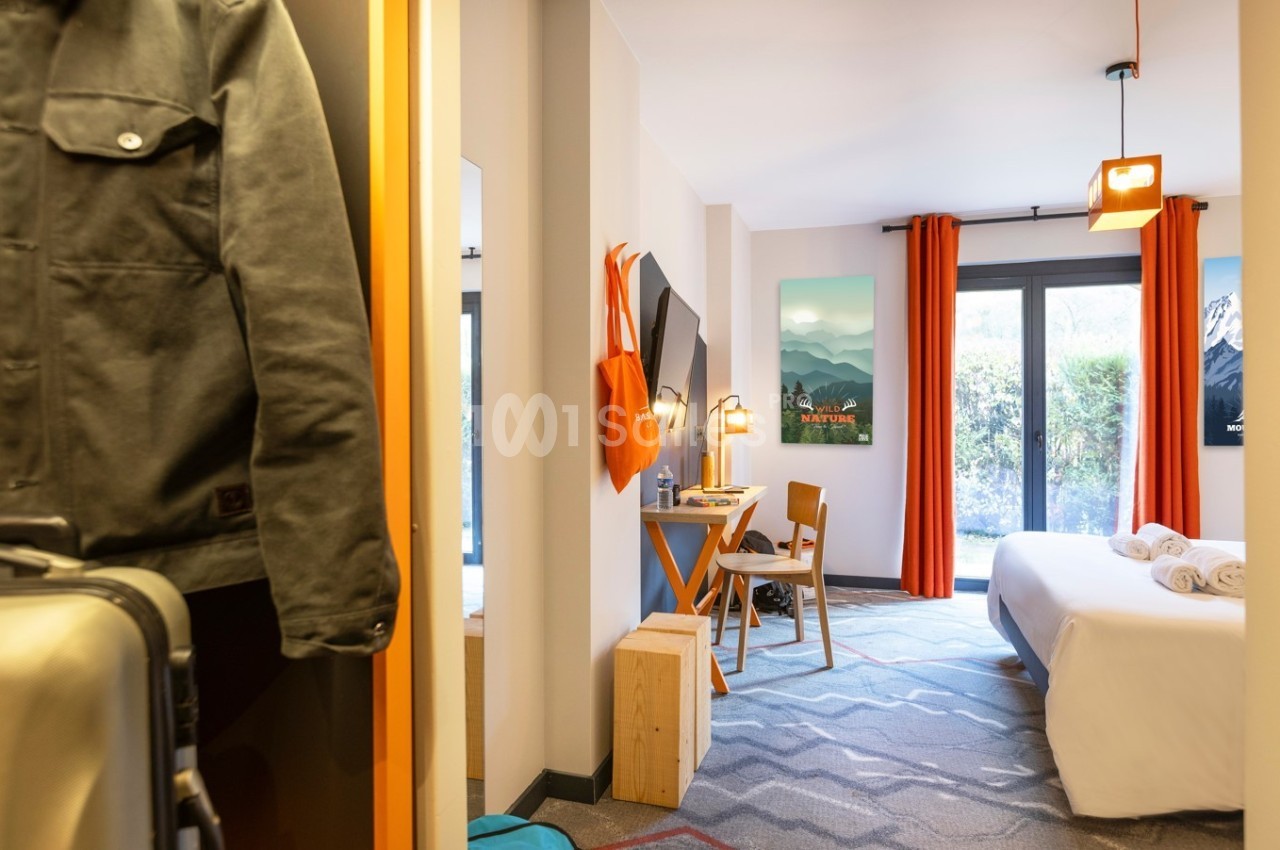 Chambre lumineuse avec lit double, bureau, rideaux orange et vue sur un jardin à travers une baie vitrée.