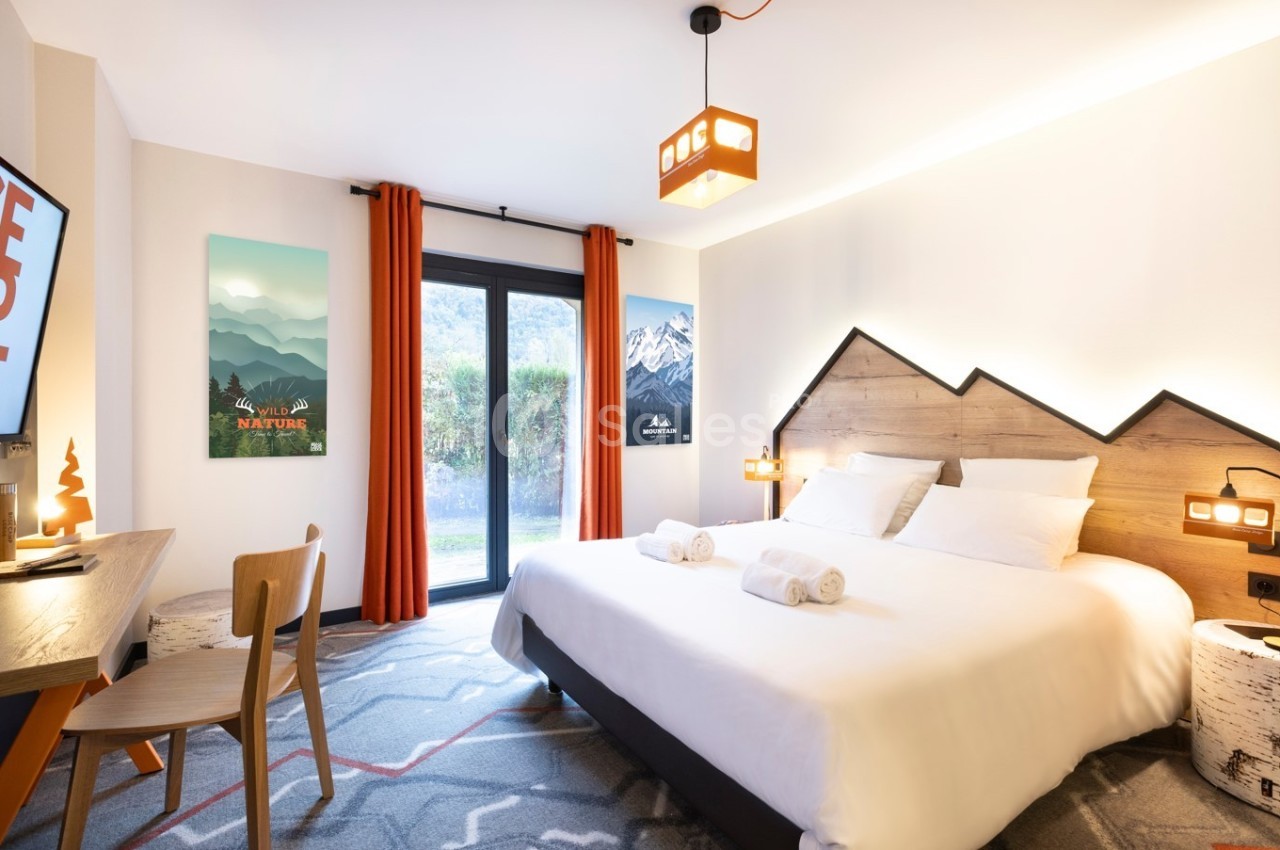 Chambre lumineuse avec lit double, décor montagnard, bureau, télévision, et vue sur un jardin par une baie vitrée.