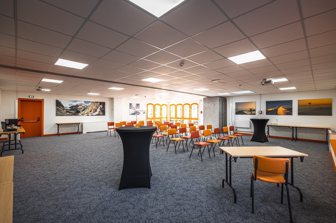 Salle de réunion lumineuse avec chaises orange disposées en rangées, tables hautes et tableaux accrochés aux murs.