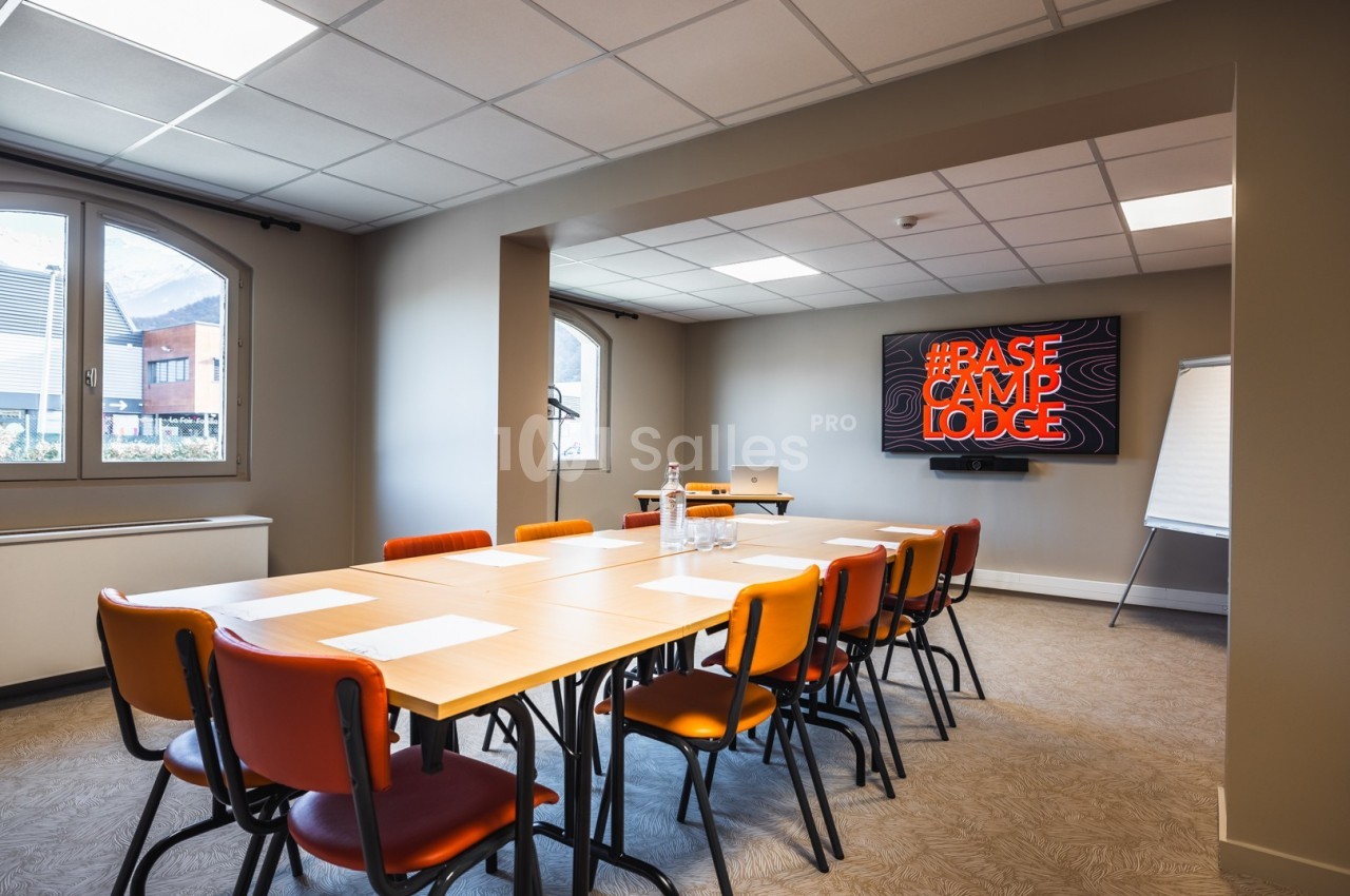 Salle de réunion lumineuse avec table rectangulaire, chaises colorées, écran affichant ’Base Camp Lodge’ et tableau blanc.