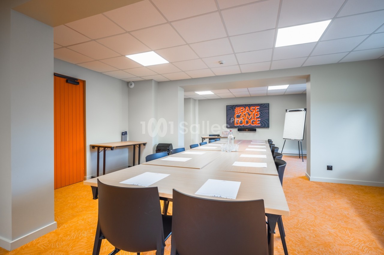 Salle de réunion lumineuse avec une grande table rectangulaire, des chaises noires et un tableau de conférence.