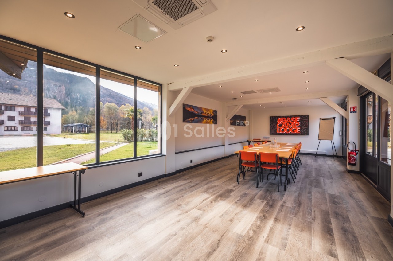 Salle de réunion lumineuse avec grandes baies vitrées, tables et chaises orange, vue sur un paysage extérieur verdoyant.