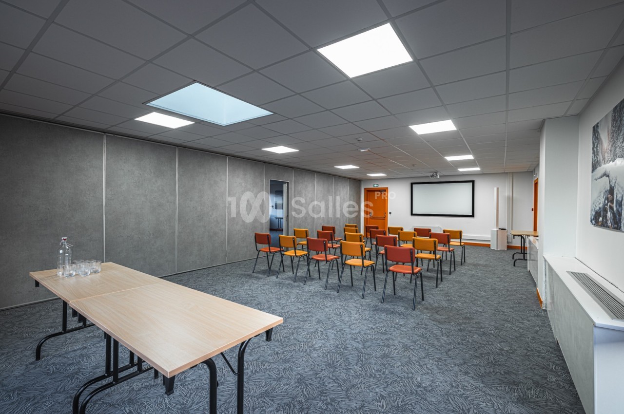 Salle de réunion avec chaises colorées alignées, écran de projection, tables en bois et éclairage au plafond.