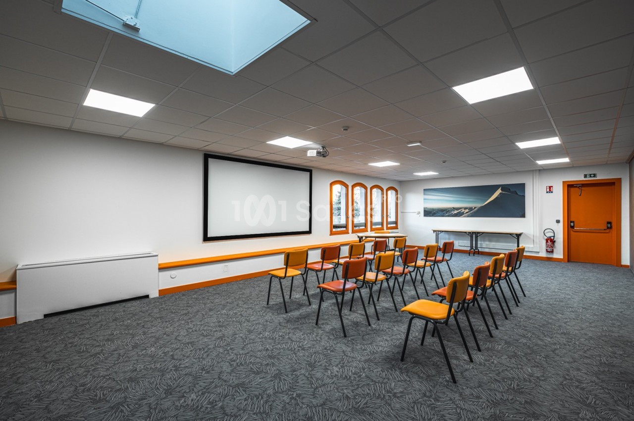 Salle de réunion lumineuse avec chaises alignées, écran de projection, fenêtres et éclairage au plafond.
