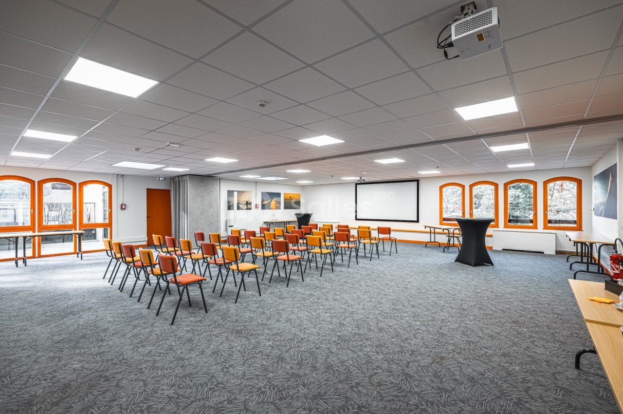 Salle de conférence lumineuse avec chaises disposées en rangées, écran de projection et grandes fenêtres.