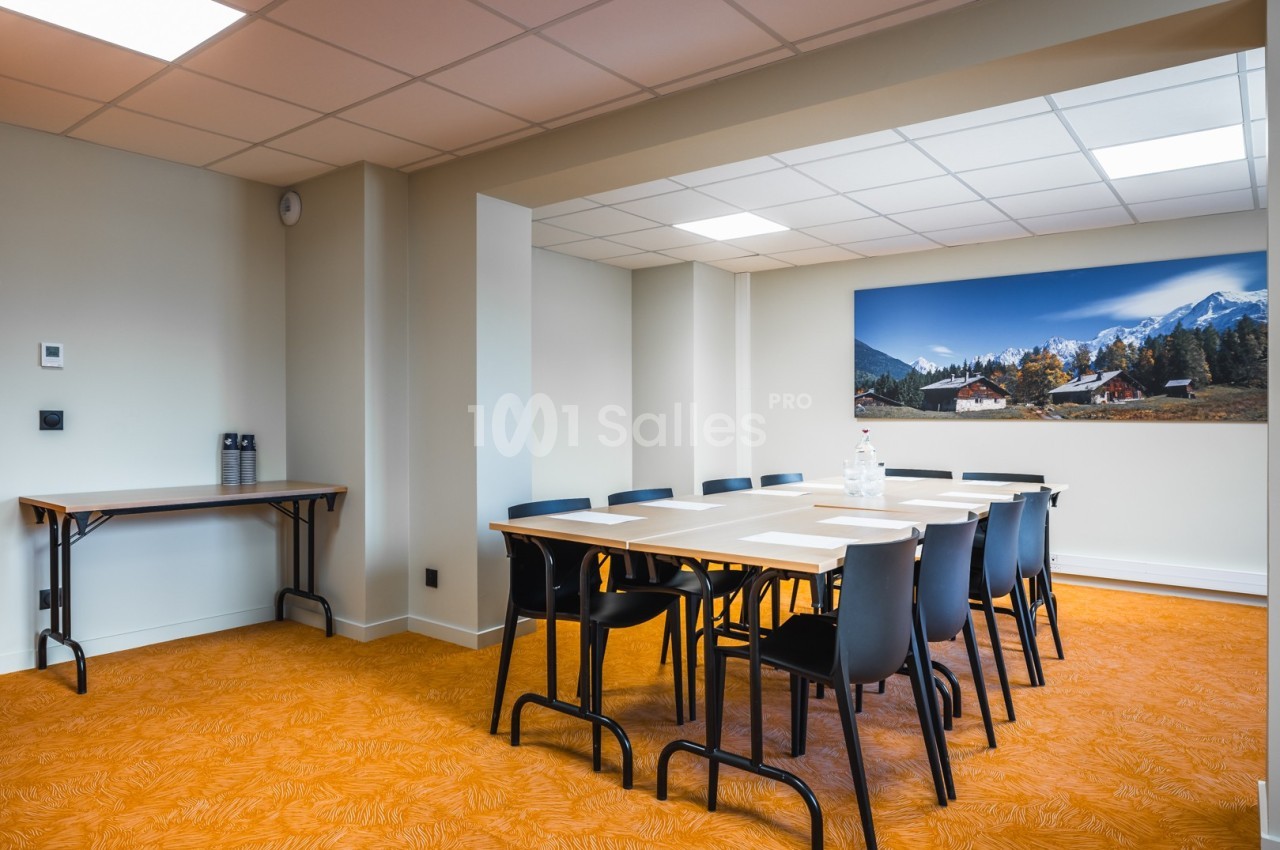 Salle de réunion lumineuse avec table rectangulaire, chaises noires, sol orange et tableau mural représentant un paysage…