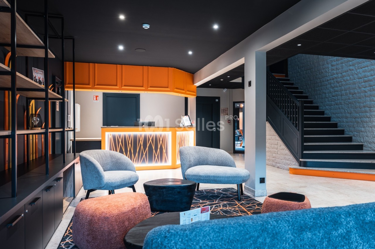 Hall d'accueil moderne avec comptoir orange, fauteuils design, étagères noires et escalier en arrière-plan.