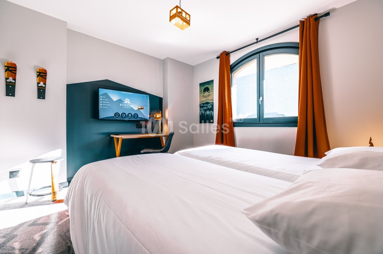 Chambre lumineuse avec deux lits simples, bureau équipé d'un écran, fenêtre avec rideaux orange et décoration murale.