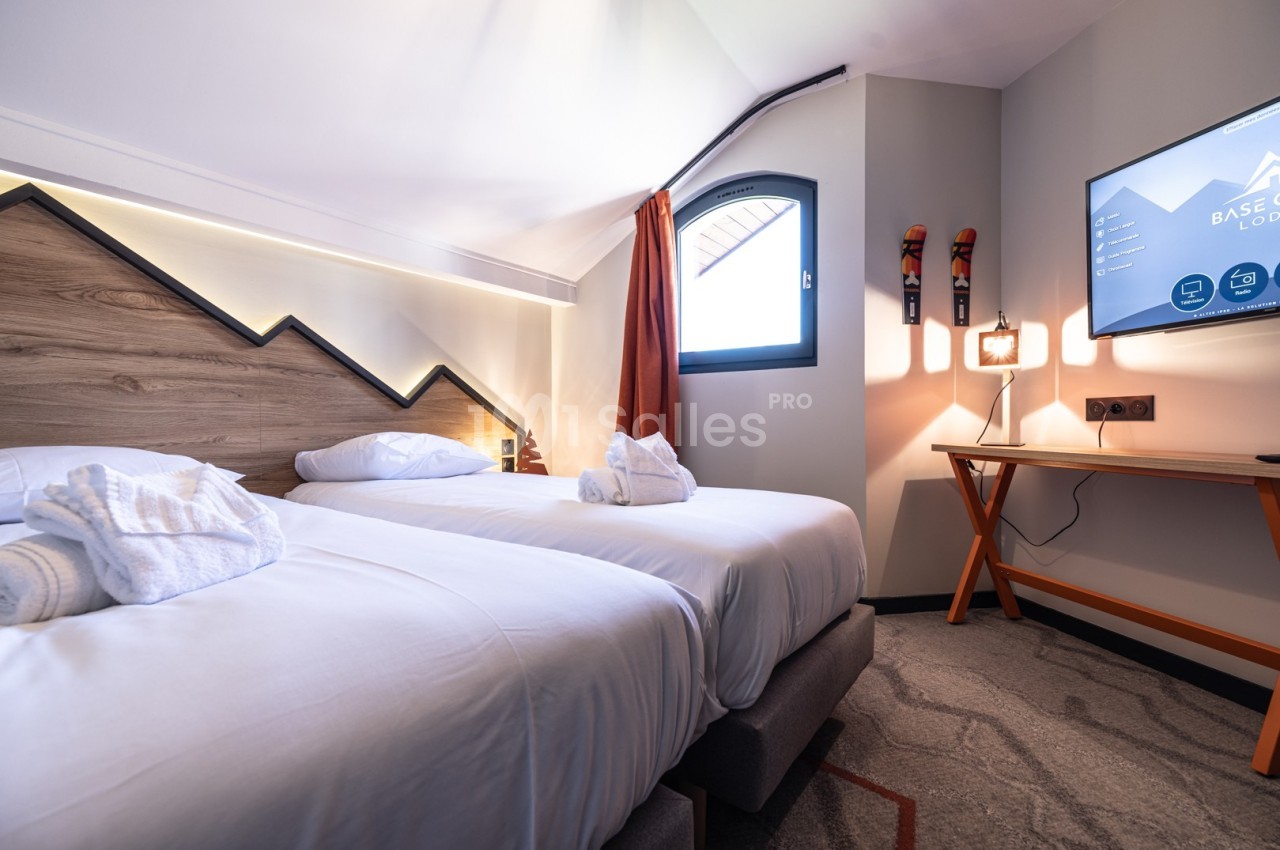 Chambre d'hôtel avec deux lits simples, linge blanc, bureau en bois, télévision murale et fenêtre avec rideau orange.
