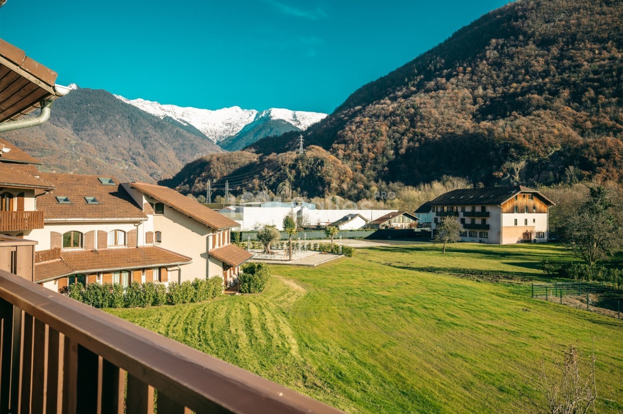 Vue depuis un balcon sur un paysage de montagne avec des maisons, des prairies et des sommets enneigés sous un ciel bleu.