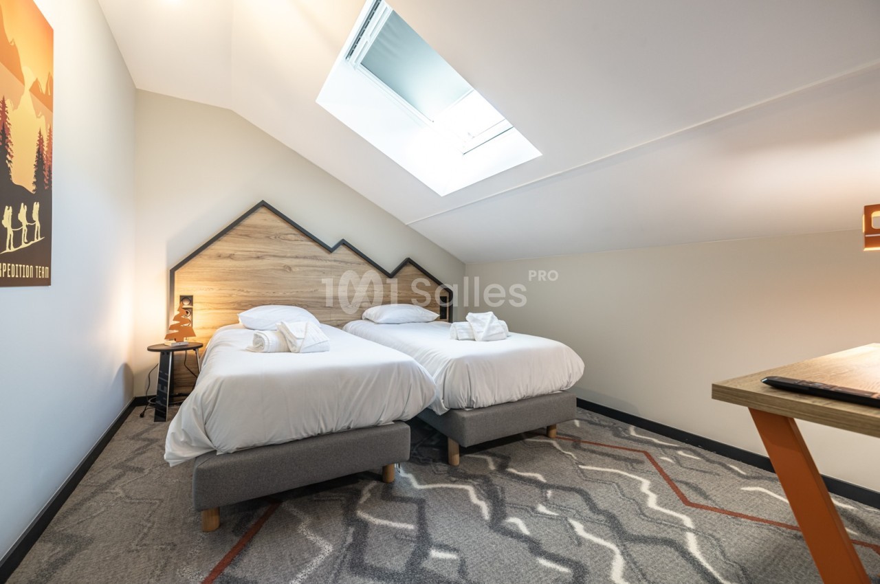 Chambre mansardée avec deux lits simples, linge blanc, éclairée par une fenêtre de toit et une lampe de chevet.