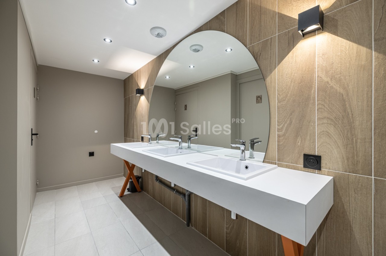Deux lavabos modernes avec miroir arrondi, éclairage intégré et mur en bois clair dans une salle de bain contemporaine.