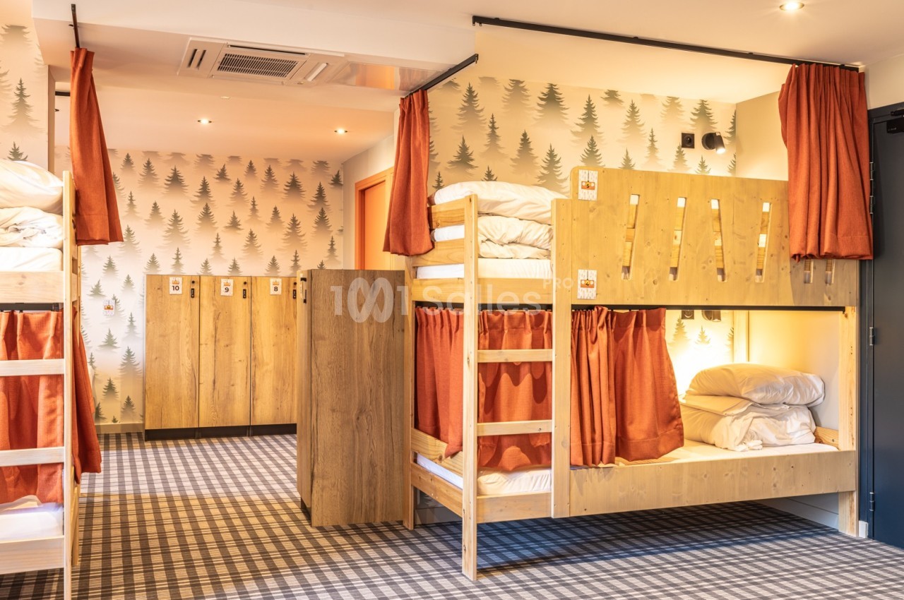 Dortoir avec lits superposés en bois, rideaux orange, casiers de rangement et papier peint à motif de sapins.