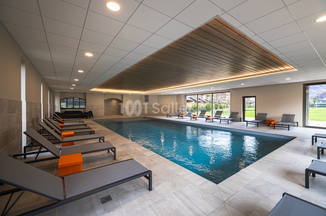 Piscine intérieure avec chaises longues grises, plafond en bois éclairé et grandes baies vitrées donnant sur un jardin.