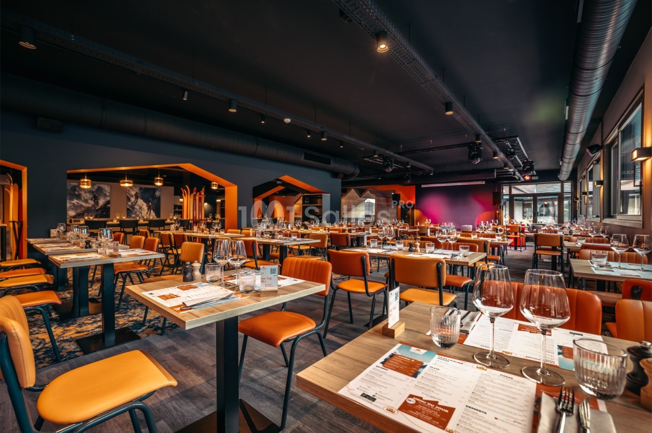 Salle de restaurant moderne avec tables dressées, chaises orange et éclairage tamisé.