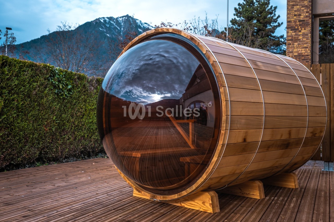 Sauna en bois en forme de tonneau avec une grande vitre, installé sur une terrasse en bois avec vue sur les montagnes.