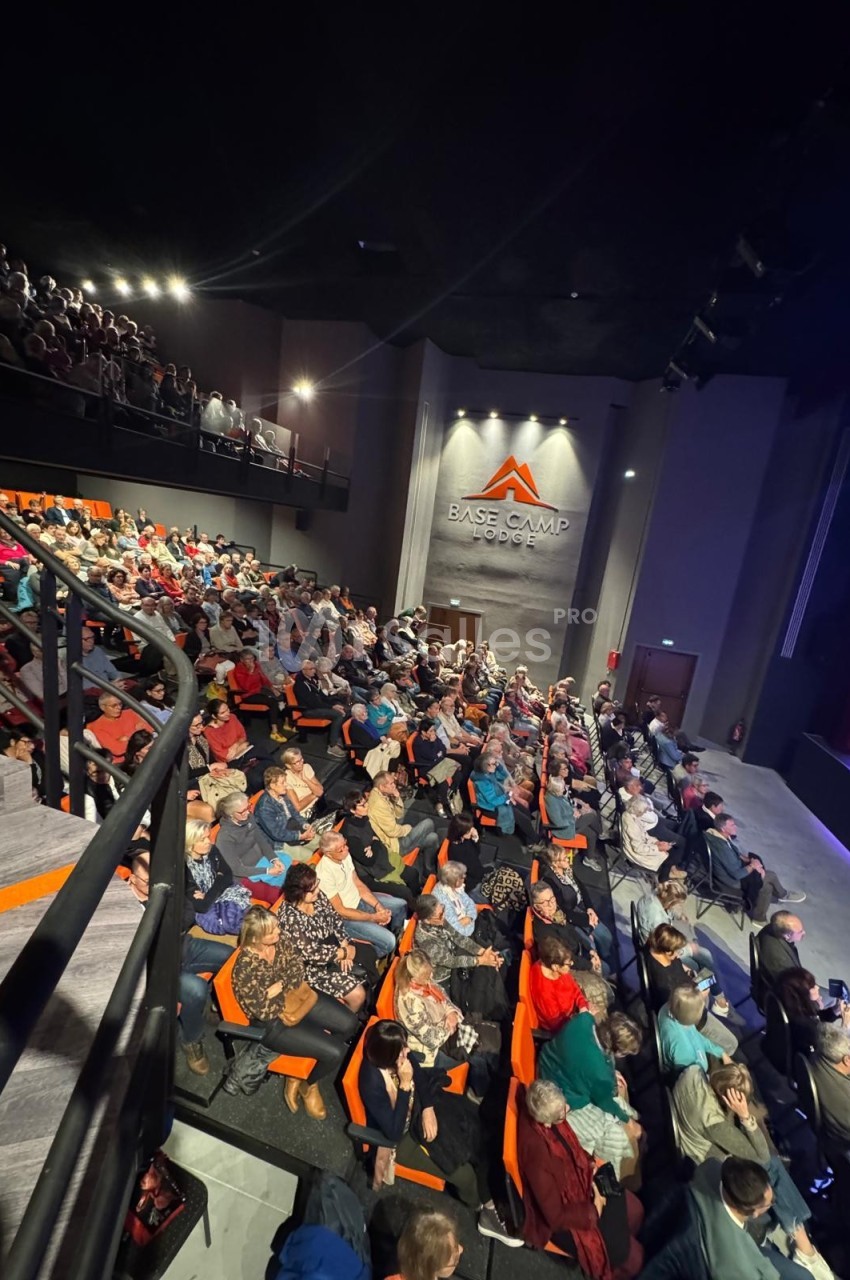 Salle de spectacle remplie de spectateurs assis sur des sièges orange, avec une scène visible sur la droite.