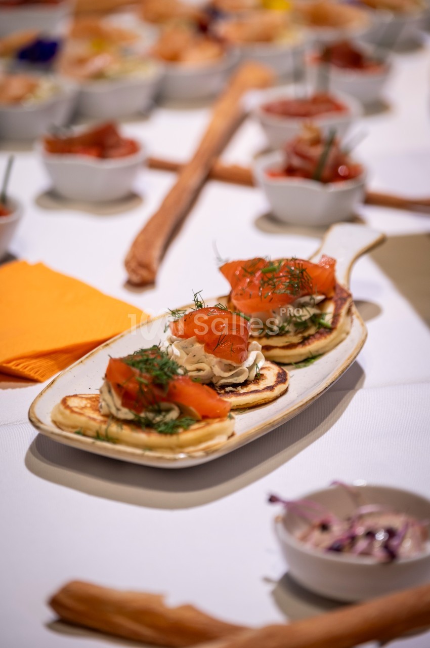 Assiette de blinis garnis de saumon fumé et crème, entourée de petites coupelles de condiments sur une table dressée.