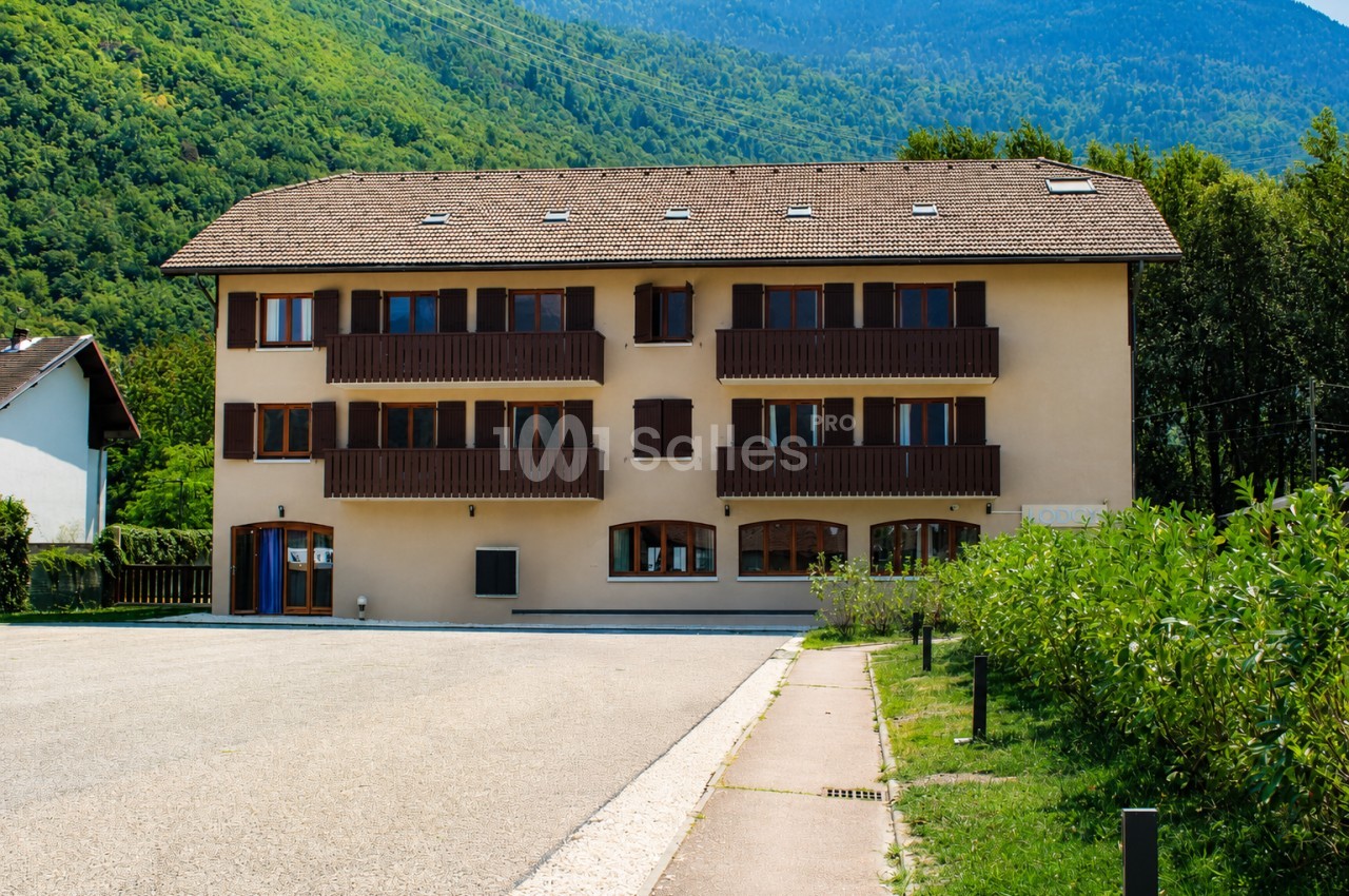 Bâtiment résidentiel à trois étages avec balcons en bois, entouré de verdure et de montagnes en arrière-plan.