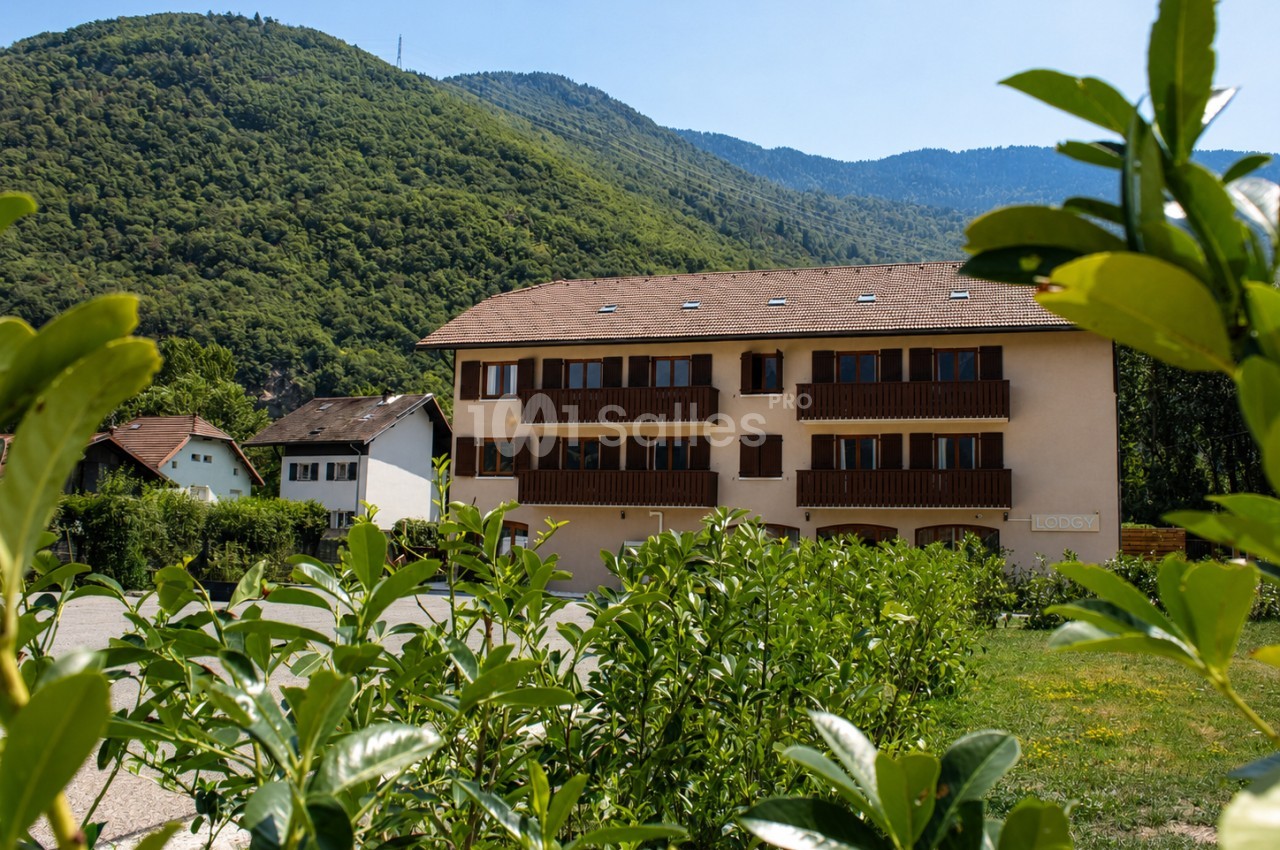 Bâtiment résidentiel beige à trois étages entouré de verdure, avec des collines boisées en arrière-plan.