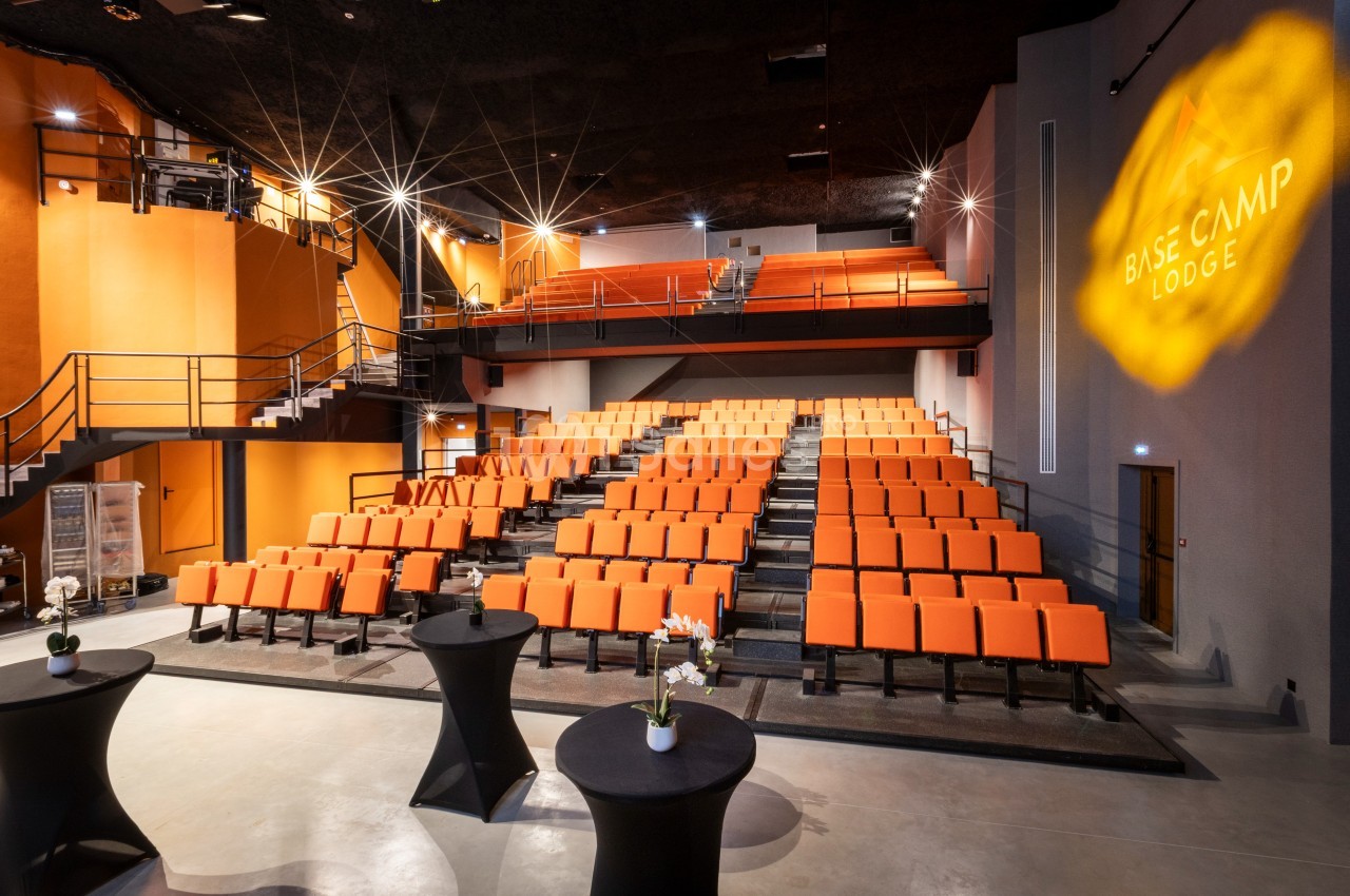 Salle de conférence moderne avec des sièges orange, un éclairage tamisé et des tables hautes au premier plan.
