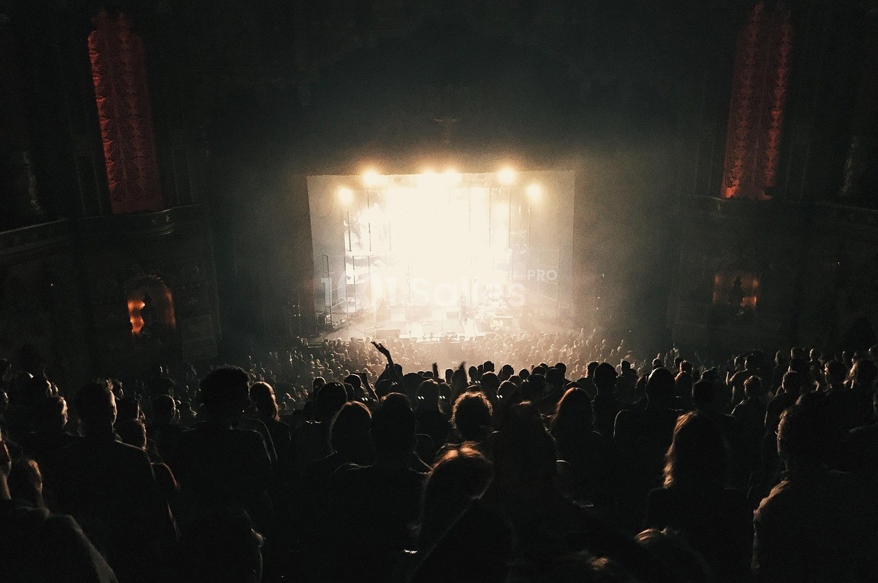 Foule dans une salle de concert sombre, éclairée par une scène lumineuse au fond.