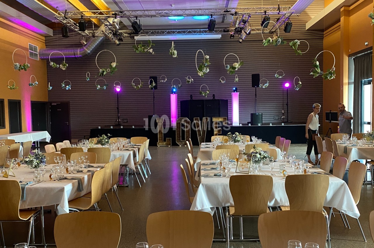 Salle de réception décorée pour un mariage, avec tables dressées, lettres ’LOVE’ lumineuses et suspensions florales.