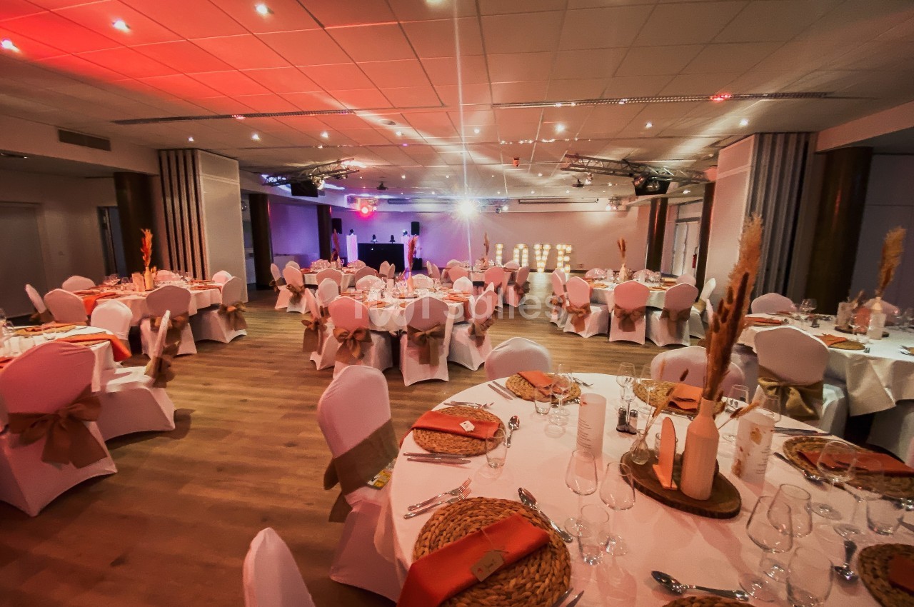 Salle de réception décorée avec des tables rondes, nappes blanches, chaises ornées de rubans et éclairage chaleureux.