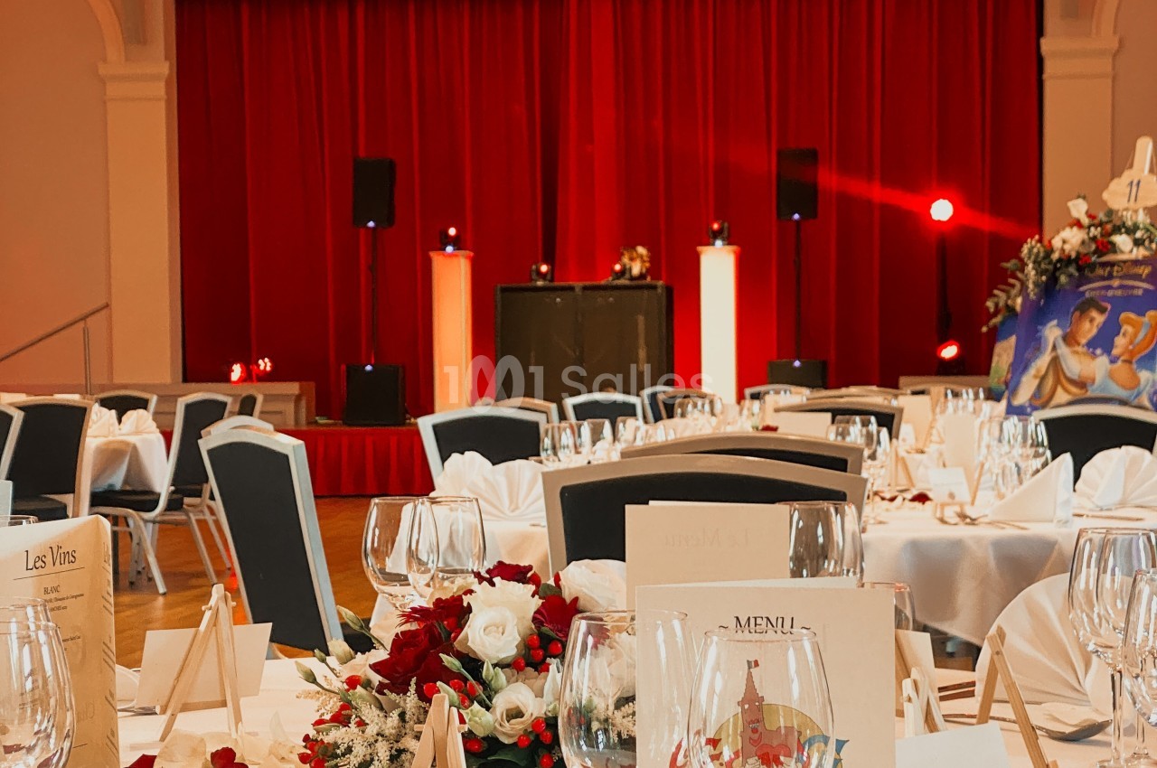 Salle de réception décorée avec des tables dressées, des fleurs et une scène avec rideaux rouges en arrière-plan.