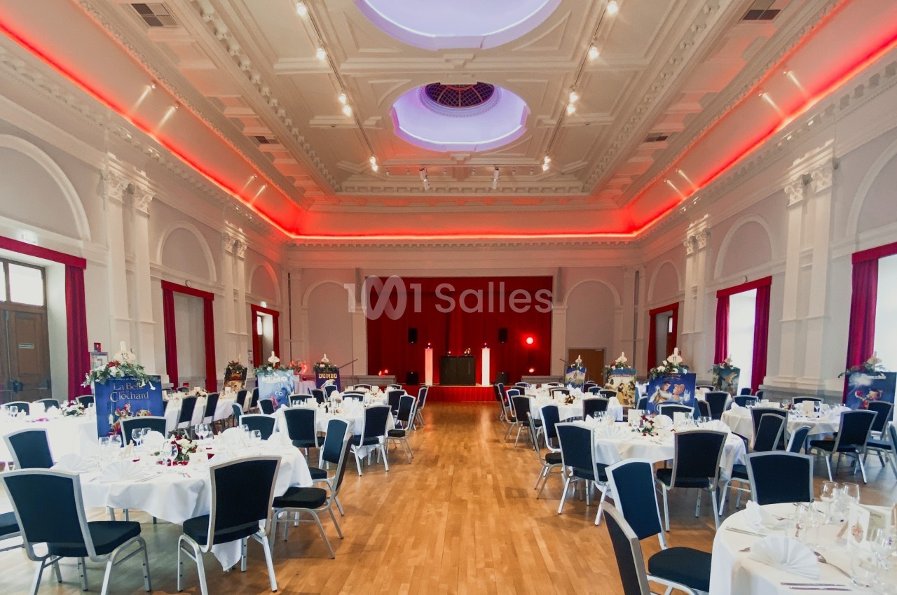 Salle de réception élégante avec tables dressées, chaises noires et rideaux rouges, éclairage tamisé au plafond.