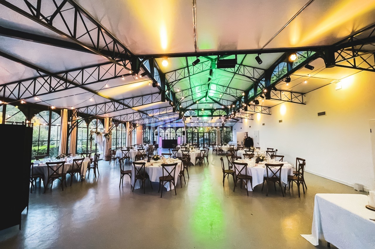 Salle de réception lumineuse avec tables rondes dressées, grandes baies vitrées et structure métallique apparente.