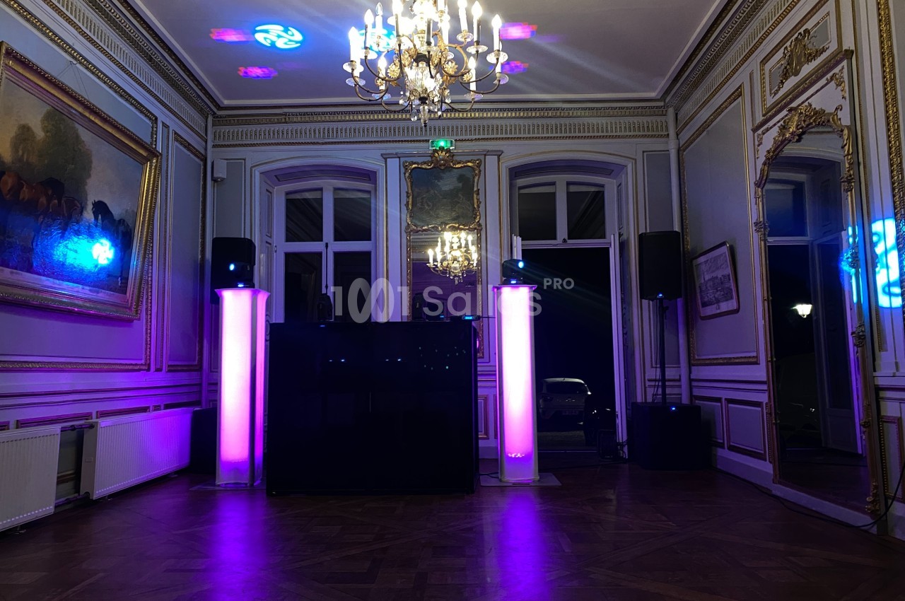 Salle élégante avec parquet, moulures dorées, lustre central et éclairage coloré autour d'une installation DJ.